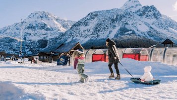 Eine Mutter zieht ihr Kind auf dem Schlitten durch den Schnee beim Familienhotel Tirolerhof, während ein weiteres Kind fröhlich vorausläuft