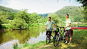 Radfahren im Thüringer Wald: Paar mit ihren Fahrrädern am Fluss Frankenoder. Im Hintergrund sind Kanus zu sehen.