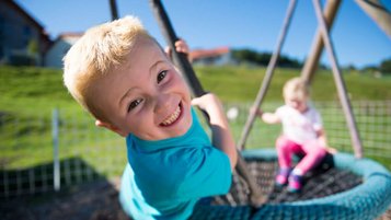 Zwei Kinder spielen im Sommerurlab im Bayerischen Wald fröhlich auf einer Nestschaukel im Garten des Familienhotels