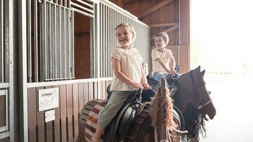 Zwei Kinder sitzen auf Holzpferden im Indoor-Spielbereich eines Familotel und lachen fröhlich.