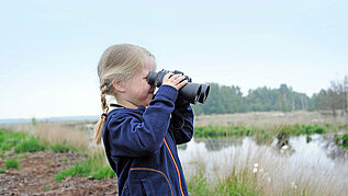 Ein junges Mädchen steht mit einem Fernglas im Dalum-Wietmarscher Moor und beobachtet die Umgebung. Im Hintergrund ist ein kleiner See zu sehen.