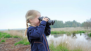 Ein junges Mädchen steht mit einem Fernglas im Dalum-Wietmarscher Moor und beobachtet die Umgebung. Im Hintergrund ist ein kleiner See zu sehen.