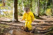 Mädchen mit gelber Matschhose und Jacke springt im Wald glücklich im Matsch herum.