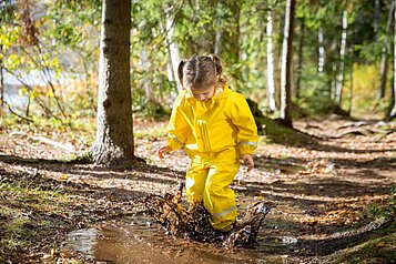 Mädchen mit gelber Matschhose und Jacke springt im Wald glücklich im Matsch herum.