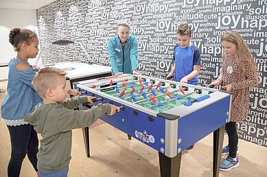 Kinder verschiedener Altersgruppen haben Spaß beim Spielen an einem farbenfrohen Tischfußball in einem Spielzimmer, dessen Wände mit Worten wie 'joy' und 'happy' dekoriert sind, was eine fröhliche und energiegeladene Atmosphäre schafft.