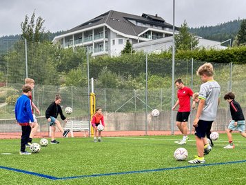 Kinder und Jugendliche trainieren mit mehreren Fußbällen auf einem Kunstrasenplatz, im Hintergrund das AIGO Hotel und bewaldete Hügel.
