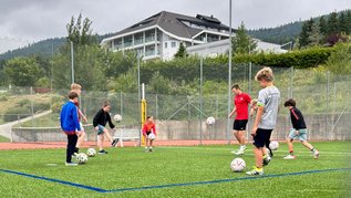 Kinder und Jugendliche trainieren mit mehreren Fußbällen auf einem Kunstrasenplatz, im Hintergrund das AIGO Hotel und bewaldete Hügel.