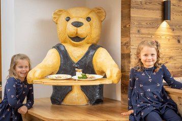 Zwei Mädchen sitzen lachend neben dem Bärenmaskottchen Bärtram mit Kindertellern im Familienhotel Kaiserhof.