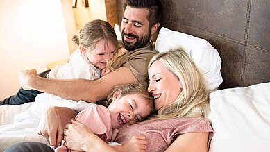 Familie mit zwei Kindern liegt im Bett eines Familienhotel und lacht mit den Kindern