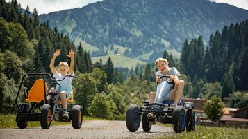 Zwei Kinder fahren lachend mit Gokarts über das Gelände des Familienhotels Krone im Allgäu, im Hintergrund die Berge.
