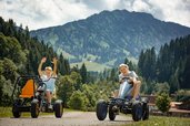 Zwei Kinder fahren lachend mit Gokarts über das Gelände des Familienhotels Krone im Allgäu, im Hintergrund die Berge.
