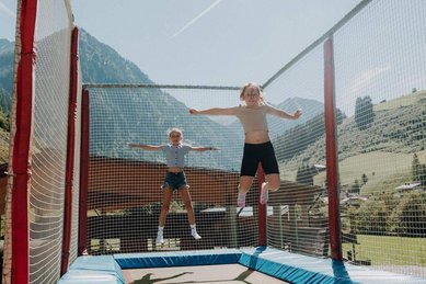 Zwei Kinder springen auf einem Trampolin im Außenbereich des Familienhotels Oberkarteis im Salzburger Land, im Hintergrund sind Berge zu sehen.