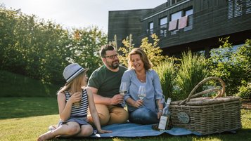 Familie sitzt mit Getränken auf einer Picknickdecke im sonnigen Garten vor einem modernen Haus, daneben ein Picknickkorb.