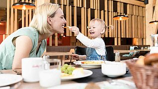 Mutter und Kind essen gemeinsam im Familienhotel. Kind füttert Mutter mit einem Stück Käse. 