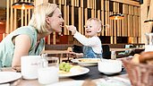 Mutter und Kind essen gemeinsam im Familienhotel. Kind füttert Mutter mit einem Stück Käse. 