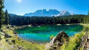 Der Karer See in Südtirol mit türkisgrünem, klarem Wasser, umgeben von dichten Nadelwäldern; dahinter schroffe Dolomiten unter blauem Himmel.