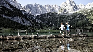 Kinder balancieren auf einem Holzstamm, dieser liegt im See in mitten einer Bergkulisse vom Familienhotel Tirolerhof an der Zugspitze.