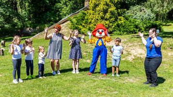 Kinderbetreuer vom Familys Club Harz spielen draußen auf der Wiese mit Maskottchen Happy und den Kindern. 