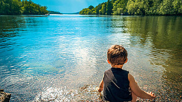 Kleinkind sitzt am Seeufer und planscht mit den Händen im Wasser.
