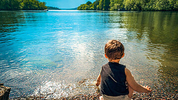 Kleinkind sitzt am Seeufer und planscht mit den Händen im Wasser.