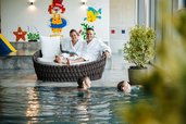 Eltern entspannen auf Relaxliegen, während Kinder im Hallenbad des Familien Resort Petschnighof in Kärnten schwimmen.