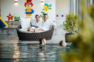 Eltern entspannen auf Relaxliegen, während Kinder im Hallenbad des Familien Resort Petschnighof in Kärnten schwimmen.