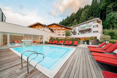 Moderner Außenpool mit roten Liegen, Holzterrasse und Blick auf die Hotelgebäude des Familienhotels Amiamo.