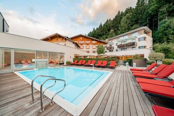 Moderner Außenpool mit roten Liegen, Holzterrasse und Blick auf die Hotelgebäude des Familienhotels Amiamo.