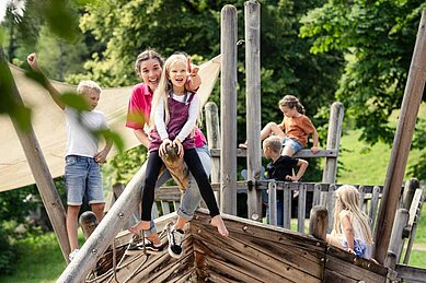 Kinder spielen gemeinsam mit einer Kinderbetreuerin auf einem Outdoor Spielplatz des Familienhotels Allgäuer Berhof.