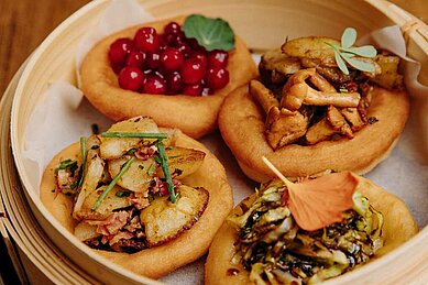 Eine Auswahl an traditionellen österreichischen Gerichten, präsentiert in einem Dampfkorb, darunter Knödel mit verschiedenen herzhaften Toppings wie Speck, Pilzen und frischen Kräutern im Hotel Habachklause.