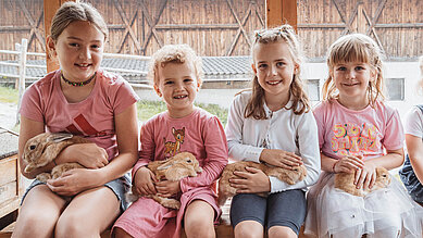 Vier Mädchen sitzen mit Hasen auf dem Schoß auf einer Fensterbank im Kleintierbereich des Familienhotels Huber in Südtirol.