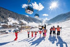 Kinder-Skikurs im Zauchenseehof mit Skilehrer und Maskottchen auf einer sonnigen Piste, im Hintergrund eine Berglandschaft mit Gondelbahn und verschneiten Hotels