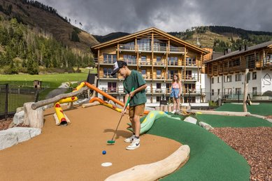 Kinder spielen Minigolf auf dem Abenteuerspielplatz des Familienhotels Almfamilyhotel Scherer mit Blick auf die umliegende Berglandschaft.