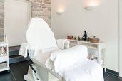 Ein heller, stilvoll eingerichteter Kosmetikraum mit weiß bezogener Behandlungsliege und Accessoires wie Klangschale und Dekorationsfigur auf einem Beistelltisch im Familienhotel Amiamo.