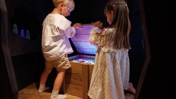 Zwei Kinder stehen vor einer beleuchteten Schatzkiste in der Magischen Abenteuerwelt des Familienhotels Kaiserhof in Tirol, aufgenommen im Bereich Feengrotte.