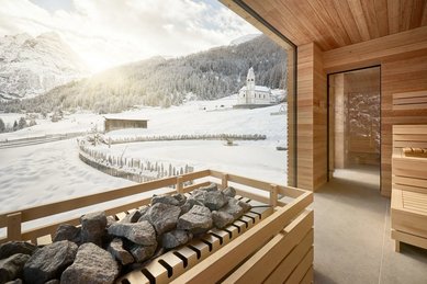 Sauna mit Aufgussbereich und Blick durch das Fenster auf eine verschneite Winterlandschaft im Hotel Bella Vista in Südtirol.