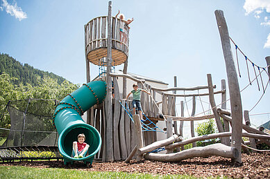 Kinder spielen auf aus dem aus Holz gebauten Outdoor-Spielplatz mit großer Röhrenrutsche im Familienhotel Huber in Südtirol.