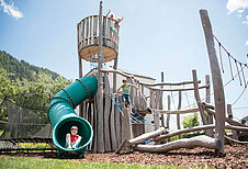 Kinder spielen auf aus dem aus Holz gebauten Outdoor-Spielplatz mit großer Röhrenrutsche im Familienhotel Huber in Südtirol.