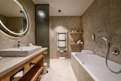 Modernes Badezimmer im Hotel Zauchenseehof mit großer Badewanne, Dusche, breitem Waschtisch mit beleuchtetem Rundspiegel, warmen Natursteinfliesen, Handtuchheizkörper und offenen Holzregalen mit neatly arrangierten Handtüchern.