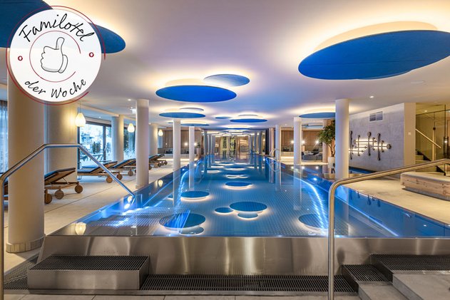 Modernes Indoor-Poolbereich im Hotel Zauchenseehof mit stimmungsvoller Beleuchtung, großen blauen Deckenpaneelen, eleganten Edelstahlbecken und gemütlichen Liegen entlang der Panoramafenster; ausgezeichnet als „Familotel der Woche“.