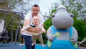 Spielplatzfreude im Familienhotel Ungarn: Ein Vater hebt sein lachendes Baby spielerisch in die Luft, neben dem beliebten Nilpferd-Maskottchen im Kolping Family Resort.