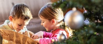 Zwei Kinder öffnen im Familienhotel Sonnenpark begeistert einen Geschenkesack neben einem festlich geschmückten Weihnachtsbaum.