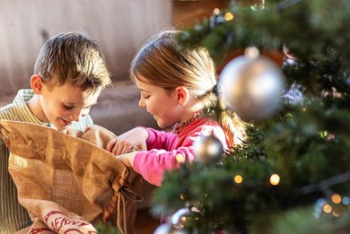 Zwei Kinder öffnen im Familienhotel Sonnenpark begeistert einen Geschenkesack neben einem festlich geschmückten Weihnachtsbaum.