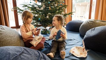 Zwei Kinder sitzen vor dem geschmückten Weihnachtsbaum im Familienhotel und packen fröhlich Geschenke aus.