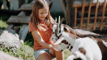Ein Mädchen sitzt im Außenbereich des Familienhotels Oberkarteis im Salzburger Land im Streichelzoo bei Ziegen und streichelt die Tiere.