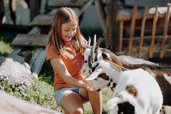 Ein Mädchen sitzt im Außenbereich des Familienhotels Oberkarteis im Salzburger Land im Streichelzoo bei Ziegen und streichelt die Tiere.