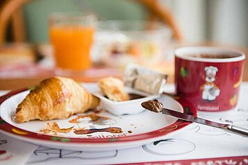 Ein leckeres Frühstück mit Croissant, Marmelade, Orangensaft und einer Tasse Kaffee auf einem Kinderplatzset im Familienhotel Sonnenhügel.