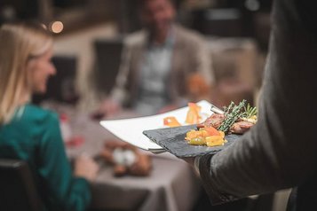 Ein Mitarbeiter bringt zwei Teller mit leckerem Essen an den Tisch im Familienhotel Alpenhof Dolomit Family.