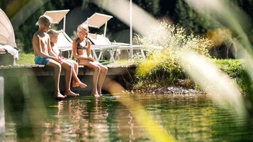 Drei Kinder sitzen auf einem Holzsteg am Naturbadeteich am Alpura Retreat in Südtirol.