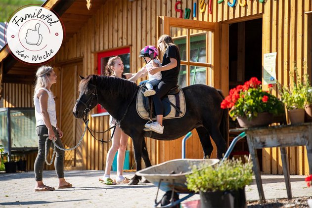Kleines Kind mit Helm sitzt auf einem schwarzen Pony, zwei Frauen helfen beim Aufsteigen vor einem Holzstall mit Blumen.